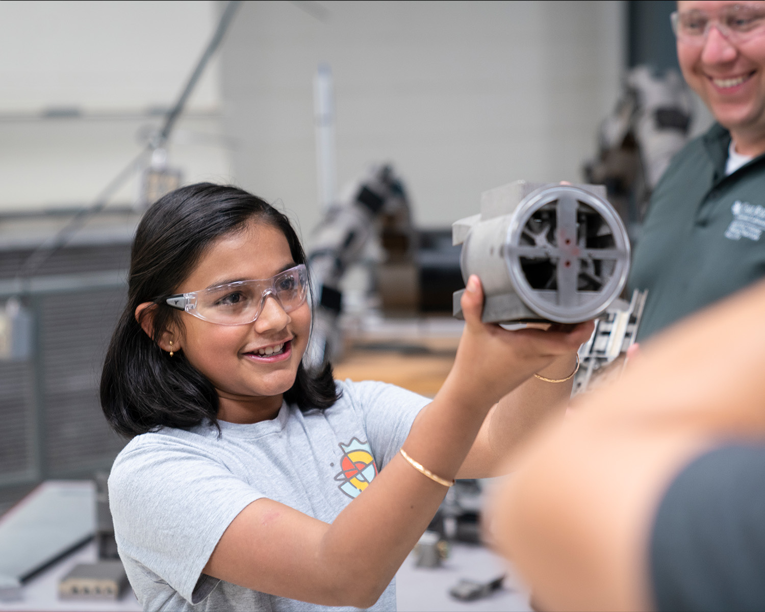 Girl wearing safety glasses and holding up mechanical component.